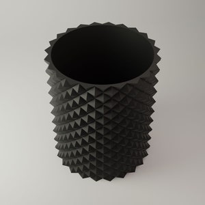 Op de afbeelding: Een zwarte cilindrische vaas met een getextureerd, stekelig exterieur. De vaas is gemaakt van een hard materiaal en heeft een gladde, ronde opening aan de bovenkant.