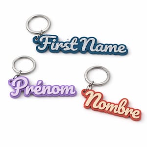 Può includere: Tre portachiavi con anelli argentati. Uno è blu con "First Name" in corsivo bianco, un altro è viola con "Prénom" in corsivo bianco, e l'ultimo è rosso con "Nombre" in corsivo crema. Tutti hanno un effetto 3D.