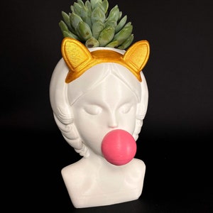 May include: White ceramic planter shaped like a woman's bust with gold cat ears and a pink bubble gum. The planter has a succulent plant growing out of the top.