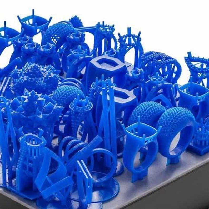 May include: A collection of blue 3D printed wax models of rings, jewelry, and other designs. The models are arranged on a gray surface.