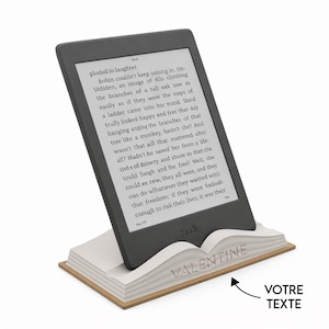 Può includere: Un e-reader nero è appoggiato su un supporto a forma di libro bianco e beige. L'e-reader mostra del testo. Il supporto ha la scritta "VALENTINE" incisa. Il supporto è realizzato in materiale chiaro.