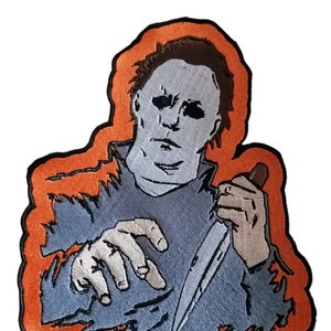 Pode incluir: Um remendo bordado com um personagem de um filme de terror. O personagem está vestindo um macacão azul e uma máscara branca. O personagem está segurando uma faca na mão direita. O remendo é cercado por uma borda laranja.