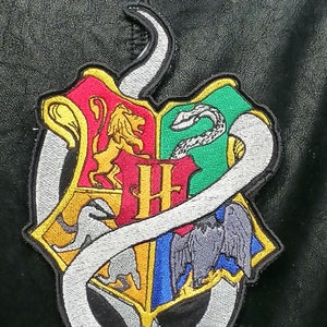 Peut inclure: Un serpent blanc avec des reflets argentés s'enroule autour d'un blason coloré avec un lion, un blaireau, un aigle et un serpent. Le blason est divisé en quatre sections, chacune avec une couleur et un animal différents. Le blason est un patch, probablement pour une veste ou un sac.