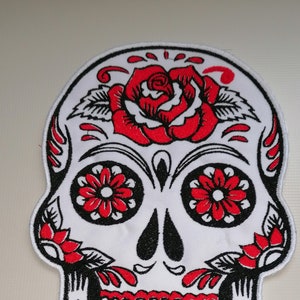 Può includere: Toppa di teschio di zucchero ricamata bianca con dettagli neri e rossi. Il teschio ha una rosa rossa sulla fronte e disegni floreali intorno agli occhi.