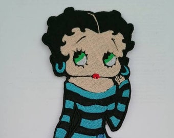 May include: An embroidered iron-on patch featuring a cartoon character with black hair, blue and black striped clothing, and blue boots. The character has large green eyes and red lips.