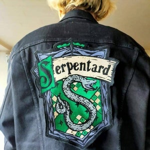 Peut inclure: Veste en jean noire avec un patch brodé vert, gris et blanc représentant un serpent et le texte "Serpentard".