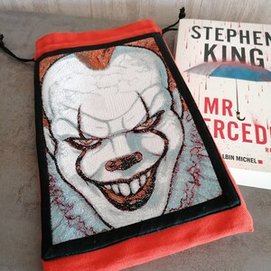 May include: A red drawstring bag with a black border and a white and red embroidered image of Pennywise the clown from the movie "It" by Stephen King. The bag is lying on a table next to a copy of the book "Mr. Mercedes" by Stephen King.