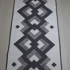 Peut inclure: Un chemin de table blanc avec un motif géométrique noir et blanc. Le motif présente une série de losanges entrelacés avec un motif zèbre.
