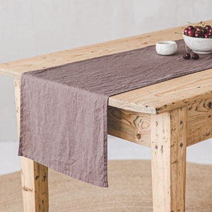 May include: A wooden table with a mauve linen table runner. A bowl of cherries and a small white bowl are on the table. The table sits on a round jute rug. The table is made of reclaimed wood.