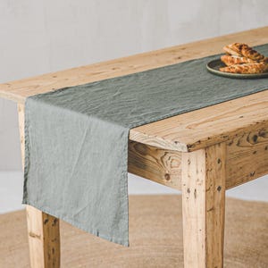 Może przedstawiać: Rustykalny drewniany stół z szałwiowym lnianym bieżnikiem. Mały talerz z wypiekami stoi na bieżniku. Stół stoi na okrągłym, tkanym dywanie jutowym.