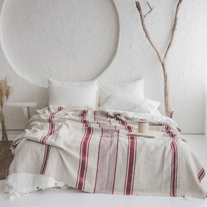 Könnte beinhalten: Ein weißes Bett mit einer rot-weiß gestreiften Wolldecke. Das Bett ist mit weißen Laken und Kissen bezogen. Im Hintergrund befindet sich eine kleine Holzbank.