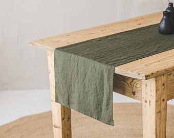 May include: A rectangular olive green linen table runner draped over a light-colored wooden table. On the runner sit two dark charcoal ceramic vases, one of which holds a single delicate stem with a small, vibrant orange-red flower petal.