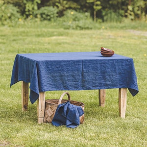 Op de afbeelding: Een blauwe linnen tafelkleed ligt op een houten tafel in een grasveld. Een rieten mand met een blauwe doek servet staat onder de tafel.