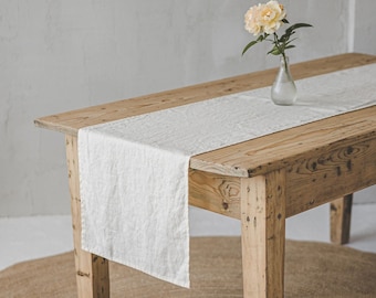 May include: A rectangular white linen table runner with a textured weave draped over a light brown wooden table. A small clear glass vase containing a single pale yellow peony flower sits on the runner, which hangs over the edge of the table.