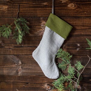 May include: A grey and green Christmas stocking hanging on a rustic wooden background. The stocking is made of linen and has a green felt cuff.