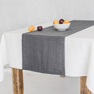 May include: A white linen tablecloth with a gray linen table runner on a wooden table. A small bowl of fruit sits on the table runner.