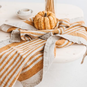 May include: A small, orange pumpkin sits on a striped linen kitchen towel. The towel is folded and draped over a wooden table. A vase of dried wheat stalks is in the background.