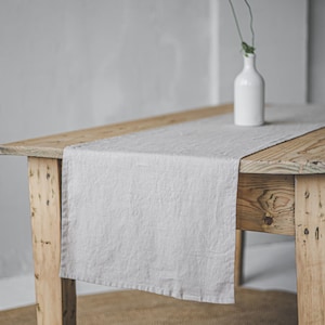 Może przedstawiać: Jasnoszary lniany bieżnik na rustykalnym drewnianym stole. Mały biały wazon zawiera pojedynczy pomarańczowy mak. Stół stoi na tkanej macie, tworząc minimalistyczną, naturalną estetykę.