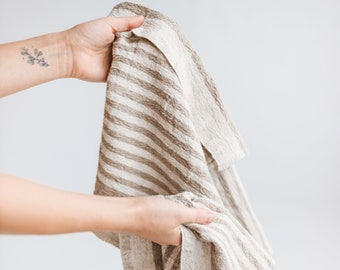 May include: A brown and white striped linen kitchen towel is held by a hand. The towel is soft and textured.