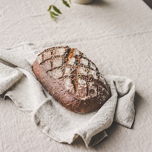 May include: A loaf of crusty bread with a diamond pattern on top, sitting on a linen napkin on a light grey table.