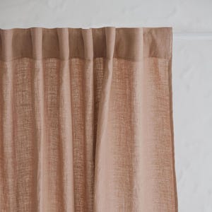 May include: A close-up of a dusty rose-coloured linen curtain. The curtain is hanging, with soft folds and a textured weave. The top of the curtain is gathered, and the fabric appears to be semi-sheer, allowing some light to pass through. The curtain is against a white wall.