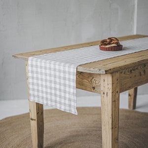 Puede incluir: Una mesa de madera rústica con un camino de mesa a cuadros gris y blanco. La mesa está puesta sobre una alfombra redonda tejida.