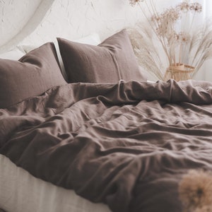 Può includere: Un copripiumino in lino marrone con due federe abbinate. La biancheria da letto è disposta su un letto con un lenzuolo bianco e una testiera in legno. Ci sono fiori secchi in un cesto di vimini sullo sfondo.