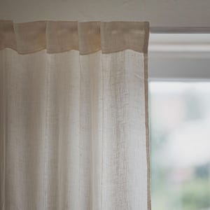 May include: Cream-coloured linen curtain panel with a textured weave. The top edge has a light beige border. The curtain hangs in soft folds, with a window visible in the background. The overall aesthetic is simple and neutral, suitable for various interior styles.