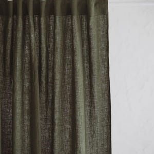 May include: Olive green linen curtain panel. The curtain is hanging, with soft folds and a textured appearance. The top of the curtain has a darker band. The background is a white wall.
