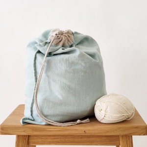 Könnte beinhalten: Eine hellblaue Leinentasche mit einem beigefarbenen Kordelzug liegt auf einem Holztisch neben einem Knäuel weißer Wolle.