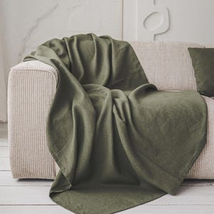 Puede incluir: Una manta de lino verde oliva sobre un sofá de pana beige. Un cojín a juego está en el sofá. El fondo presenta una pared blanca y una obra de arte abstracta enmarcada. La manta añade un toque de color y textura al espacio habitable de tonos neutros.