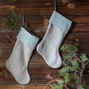 May include: Two grey linen Christmas stockings with blue felt cuffs hanging on a brown wooden background.  Green pine branches are scattered around the stockings.