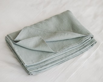 Puede incluir: Una pieza rectangular de tela color verde salvia doblada con una textura tejida visible y bordes cosidos. El textil está dispuesto en varios pliegues ordenados, mostrando un tono verde suave y apagado en toda la superficie del material.