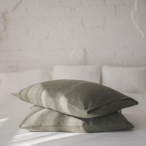 May include: Two green linen pillowcases stacked on a white bed. The pillowcases are made of a soft, natural fabric and have a subtle texture.