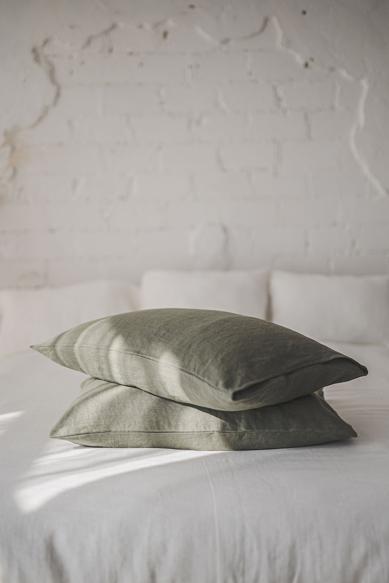 May include: Two green linen pillowcases stacked on a white bed. The pillowcases are made of a soft, natural fabric and have a subtle texture.