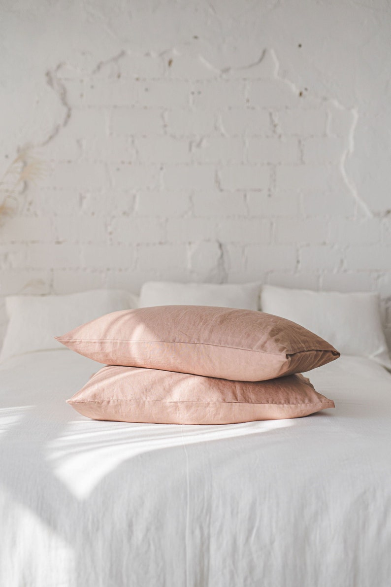 May include: Two blush pink linen pillowcases stacked on a white bed. The pillowcases are made of a soft, natural fabric and have a subtle sheen.