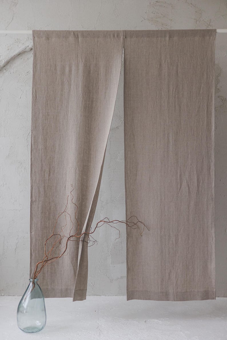 May include: A pair of beige linen curtains hanging from a white rod. The curtains are open in the middle, revealing a white wall behind them. A dried branch in a clear glass vase sits in front of the curtains.