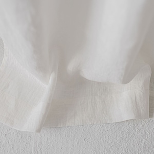 May include: A white fabric with a visible woven texture and a straight hemline. The material is draped in soft folds, highlighting the natural weave and the semi-sheer quality of the textile. The edges are finished with a clean, stitched hem.