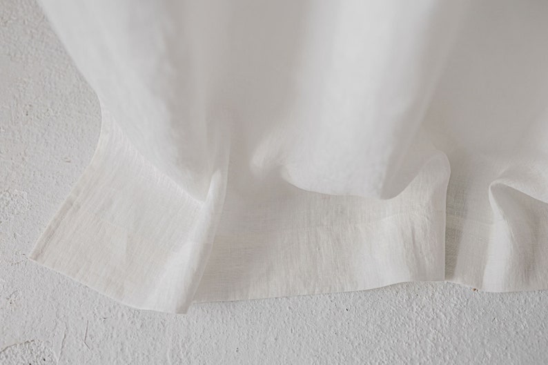 May include: A white fabric with a visible woven texture and a straight hemline. The material is draped in soft folds, highlighting the natural weave and the semi-sheer quality of the textile. The edges are finished with a clean, stitched hem.
