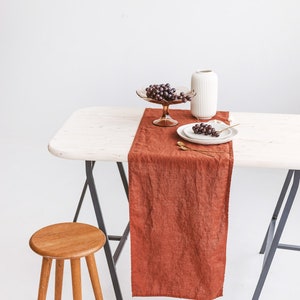 May include: A rustic table setting with a brown linen table runner, a white vase, a glass bowl of grapes, and two white plates with gold silverware. The table is on a pair of black sawhorses and a wooden stool is in the foreground.
