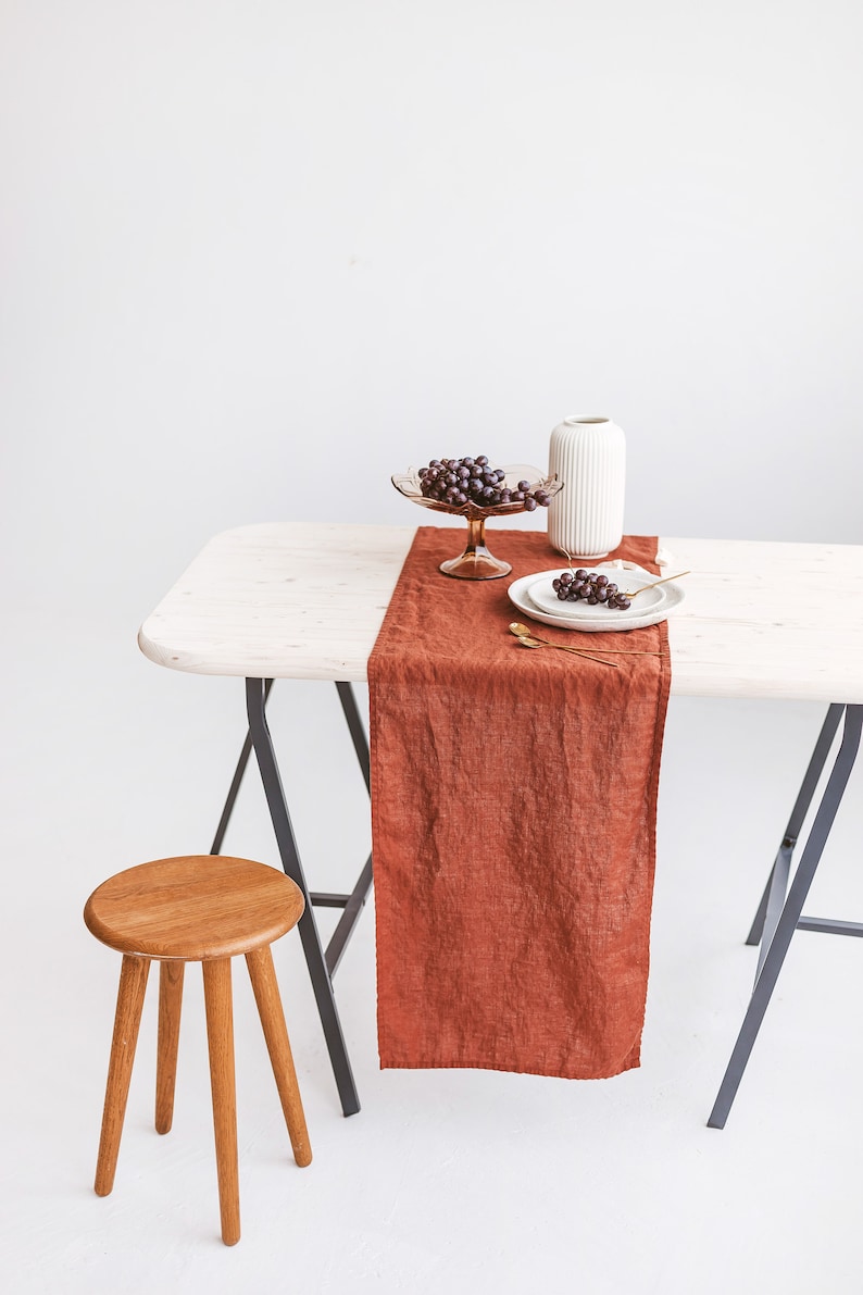 May include: A rustic table setting with a brown linen table runner, a white vase, a glass bowl of grapes, and two white plates with gold silverware. The table is on a pair of black sawhorses and a wooden stool is in the foreground.