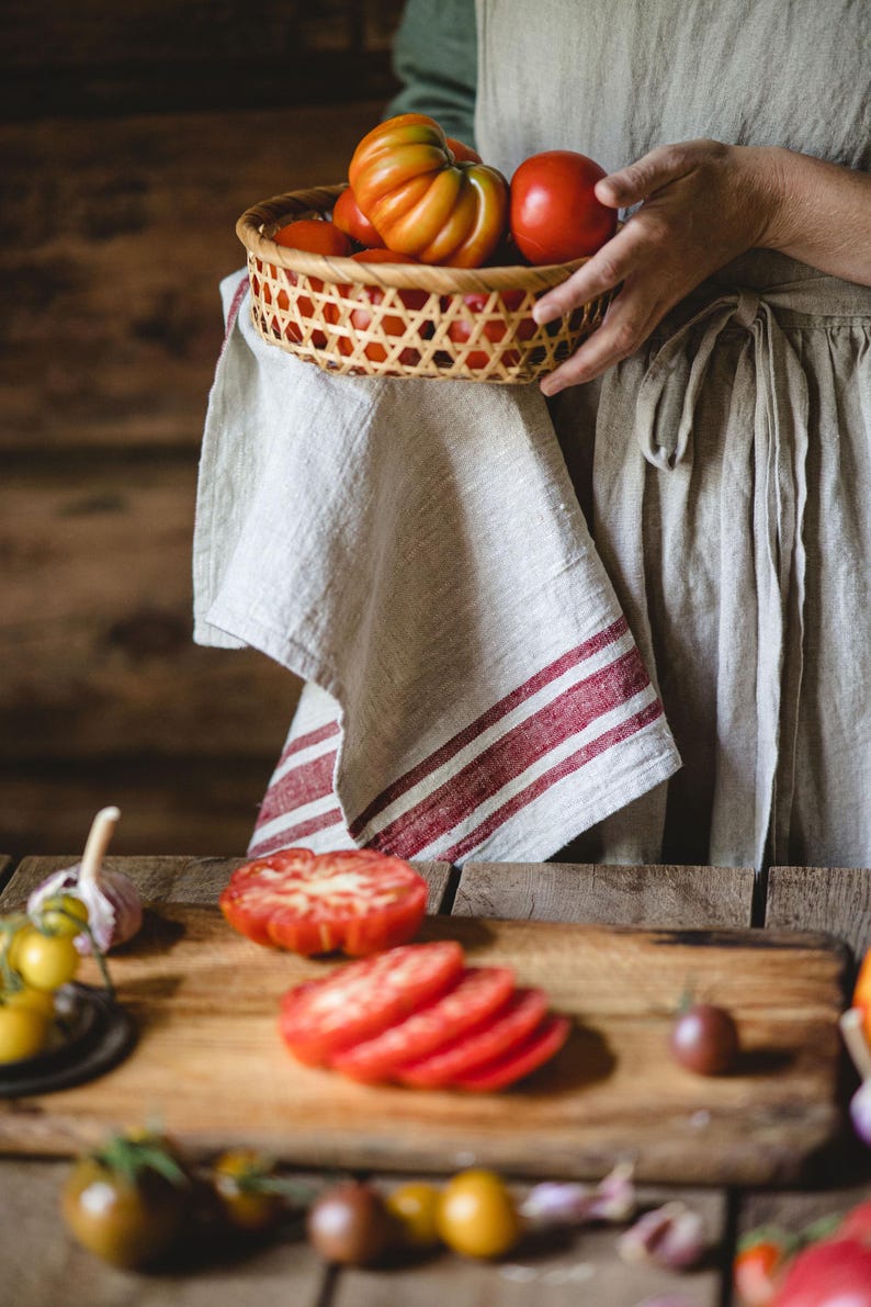 Puede incluir: Una persona sosteniendo una cesta tejida llena de tomates rojos y naranjas. Una tabla de cortar de madera con tomates en rodajas y otras verduras est&aacute; sobre una mesa de madera.