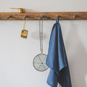 May include: A rustic wooden wall rack with multiple pegs holds a gold-toned metal measuring cup, a wire mesh skimmer with a long handle, and a deep blue linen kitchen towel hanging from a fabric loop, all arranged neatly in a row.
