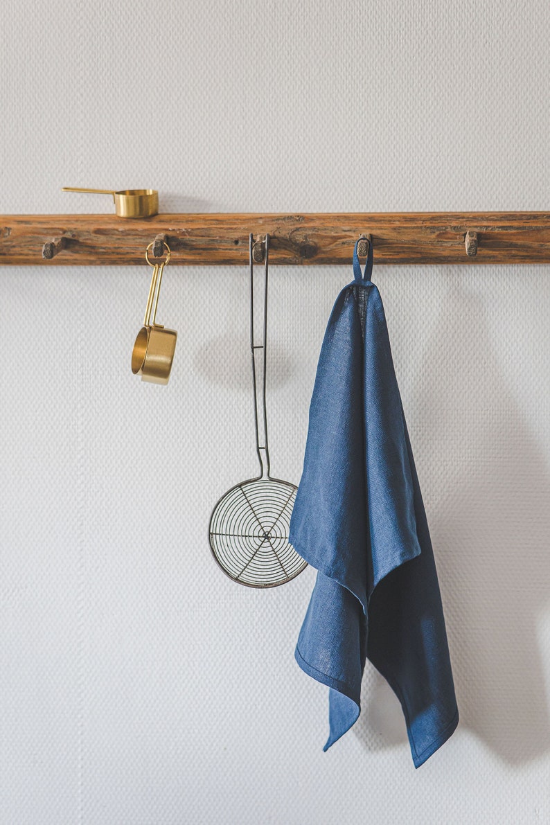 May include: A rustic wooden wall rack with multiple pegs holds a gold-toned metal measuring cup, a wire mesh skimmer with a long handle, and a deep blue linen kitchen towel hanging from a fabric loop, all arranged neatly in a row.