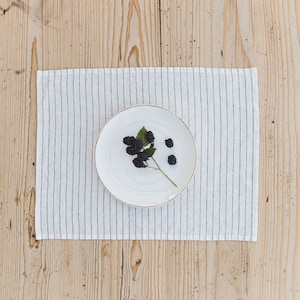 May include: A white plate with a gold rim holds fresh blackberries and a green leaf. The plate rests on a white and black striped placemat. The placemat and plate are on a light-coloured wooden table.
