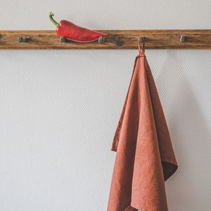 May include: A rustic wooden wall rack with multiple pegs holds a single bright red chili pepper on the left side and a solid terracotta-colored linen kitchen towel hanging from a central loop on the right side against a textured white wall.