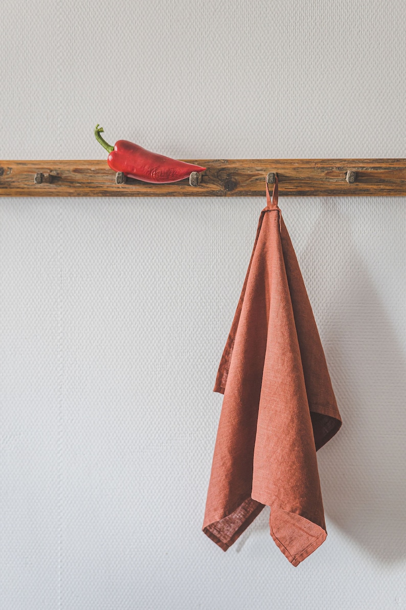 May include: A rustic wooden wall rack with multiple pegs holds a single bright red chili pepper on the left side and a solid terracotta-colored linen kitchen towel hanging from a central loop on the right side against a textured white wall.