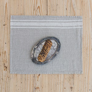 May include: A gray linen placemat with white stripes, featuring a small oval-shaped plate with a brown and black breadstick on top.