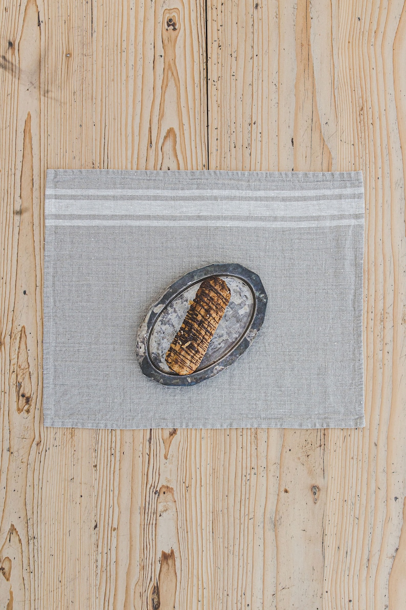 May include: A gray linen placemat with white stripes, featuring a small oval-shaped plate with a brown and black breadstick on top.