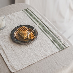 May include: A piece of layered cake with a brown and white filling on a silver plate with a spoon. The plate is on a beige linen tablecloth with green stripes.